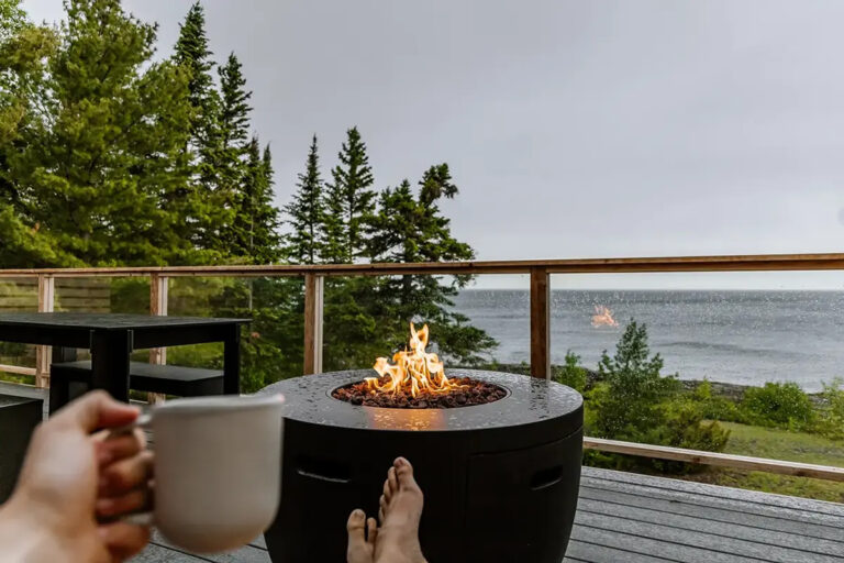 A person enjoying coffee in front of a fire pit and an epic lake view at Fresh Coast Cabins.